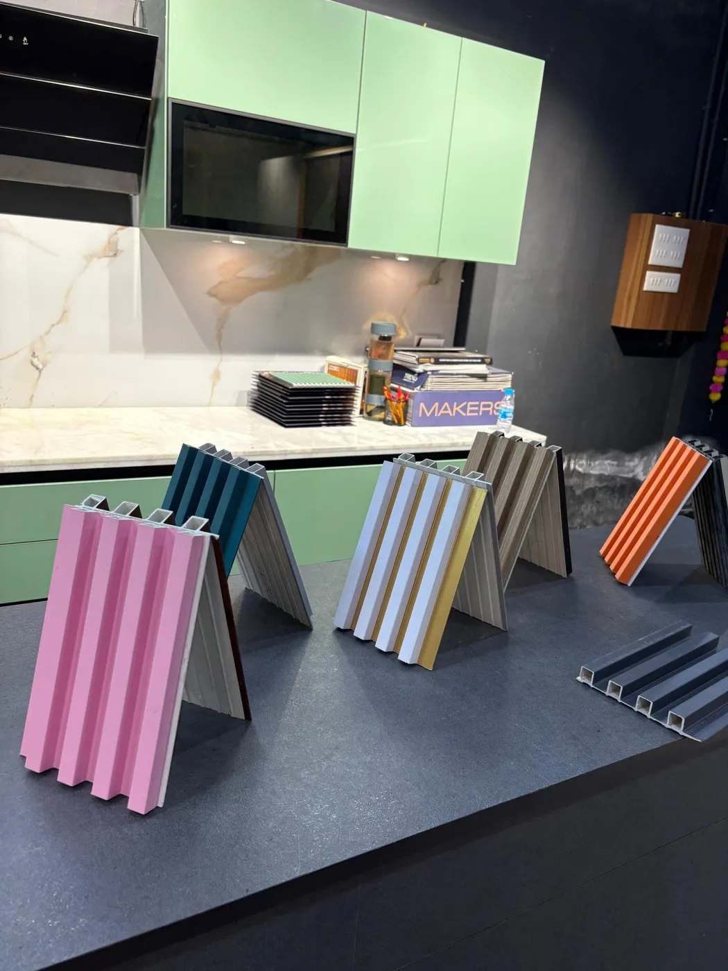 Hollow-profile fluted louver samples in pink, teal, white-and-gold, wood and orange, standing on a kitchen counter